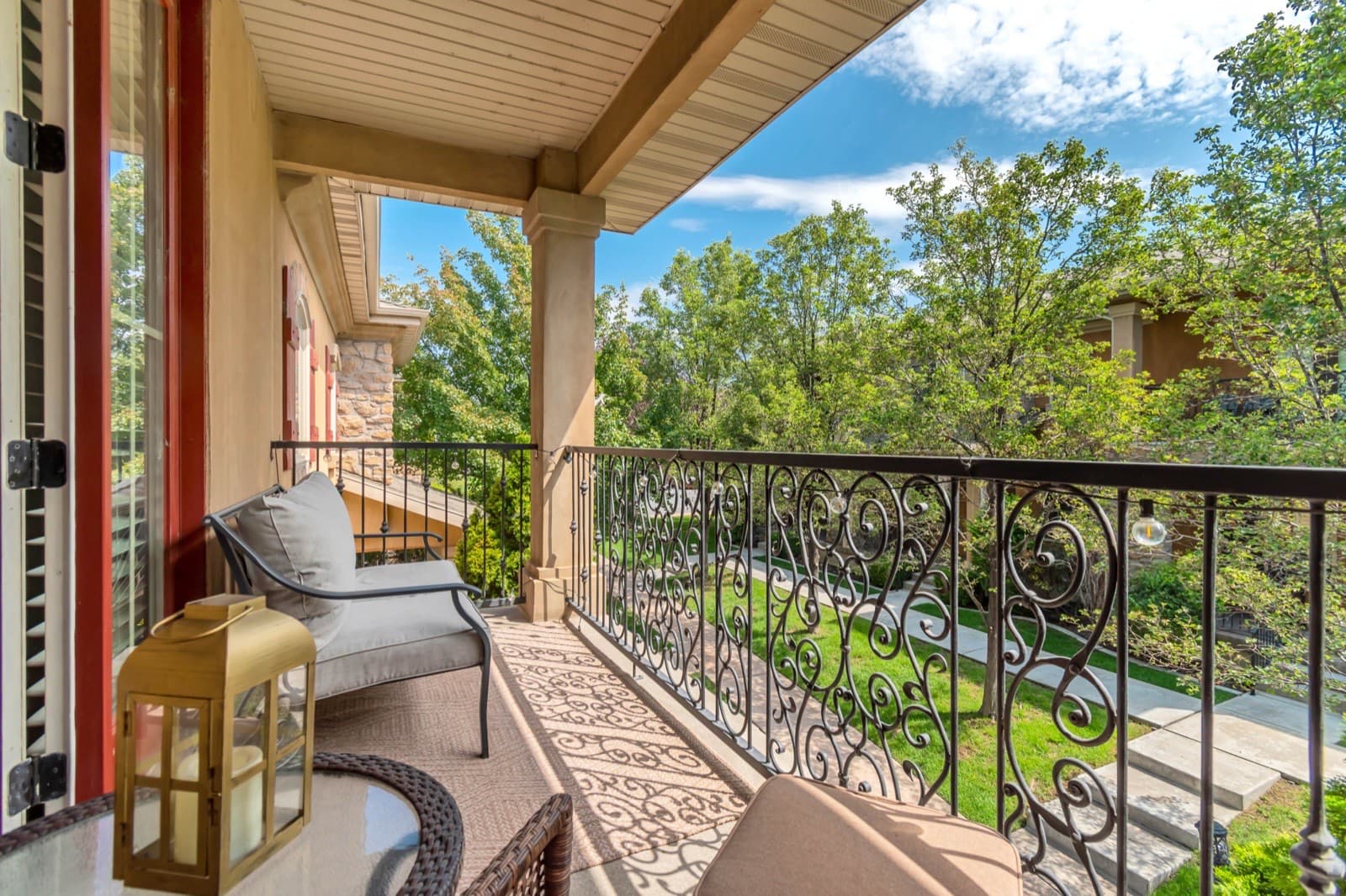 Private covered balcony off the primary suite with a wrought-iron rail, stamped floor, and seating under the tree canopy